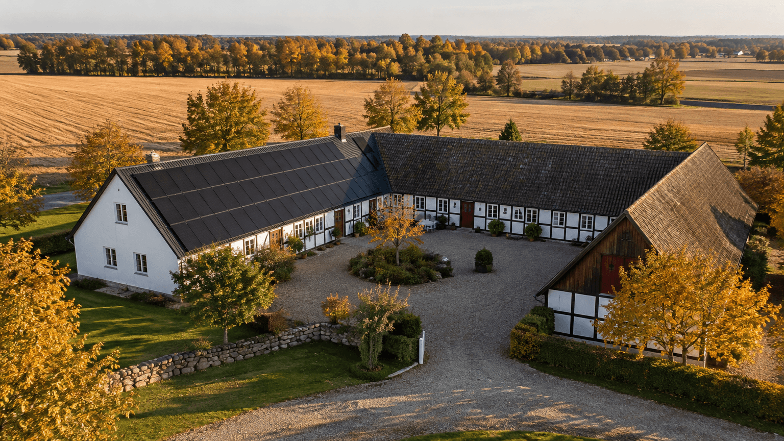 Flygbild över en vitkalkad skånsk gård med korsvirke och svarta solpaneler på ena längans tak bland gyllene höståkrar