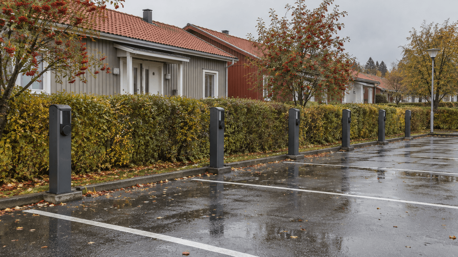 Rad med mörkgrå laddstolpar vid parkeringsplatser framför låga radhus med rött tegeltak, rönnbär och våt asfalt en mulen höstdag